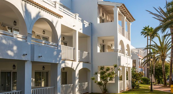 A white Mediterranean-style building with arches, balconies, and outdoor lighting, surrounded by palm trees and greenery under a clear blue sky.