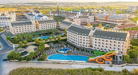 Large resort complex with multiple connected buildings, surrounded by green gardens and pools. Features include a large main pool with slides and an amphitheater area. The architecture blends modern design with classic touches, giving a luxurious and welcoming atmosphere.