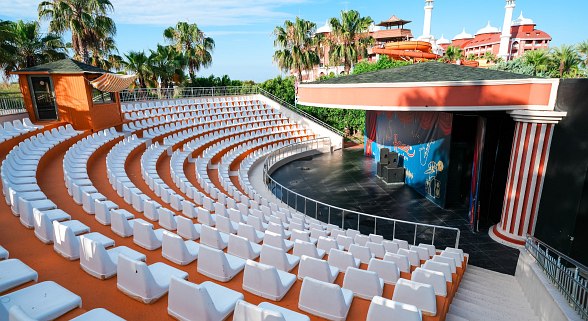 Outdoor amphitheater with rows of white seats arranged in a semi-circle around a stage. The stage has a roof and decorative columns, with a colorful backdrop. Surrounded by palm trees and buildings, it looks like a venue for live performances or shows in a warm climate.