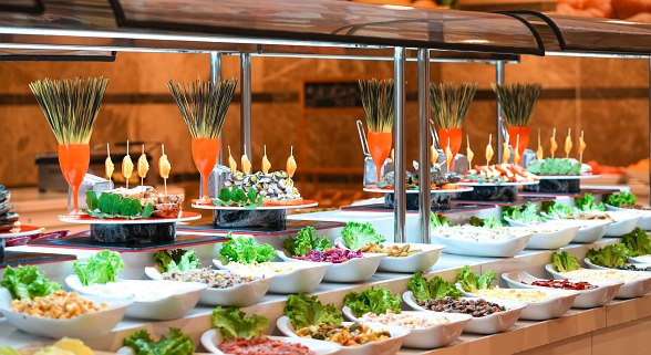 Buffet spread with various dishes presented beautifully. Multiple plates with appetizers and salads are arranged neatly on a long counter. The setup is colorful and inviting, ideal for a diverse dining experience.