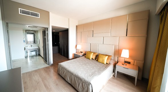 A modern hotel room with a large bed, beige bedding, gold accent pillows, bedside tables with lamps, wooden flooring, and a view into a bright bathroom through glass doors.