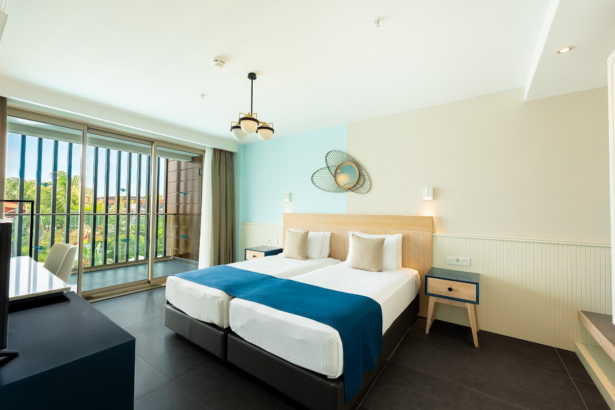 Modern hotel room with two single beds joined, white linens, beige and blue bedding, a window with sheer curtains, a vanity with a round mirror, and a TV on a wooden stand.