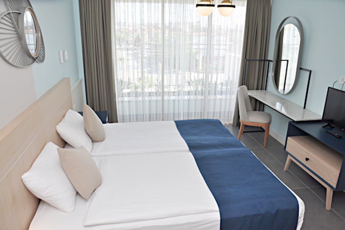Modern hotel room with two single beds joined, white linens, beige and blue bedding, a window with sheer curtains, a vanity with a round mirror, and a TV on a wooden stand.