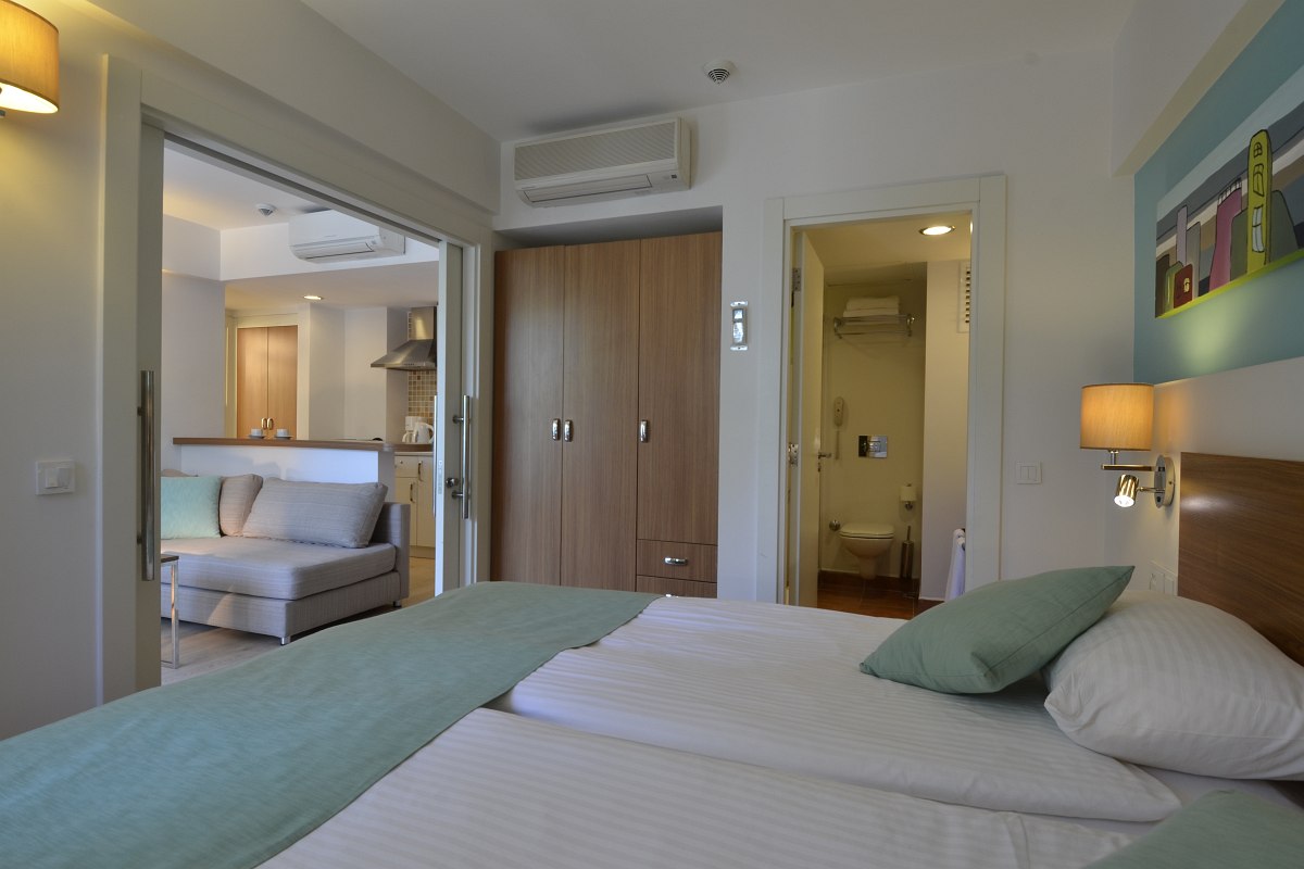 Cozy hotel room with twin beds, green bedding, a wooden headboard, and wall-mounted lights. The room opens to a living area with a sofa and kitchen. There's an air conditioner above the wardrobe and a bathroom visible through an open door.