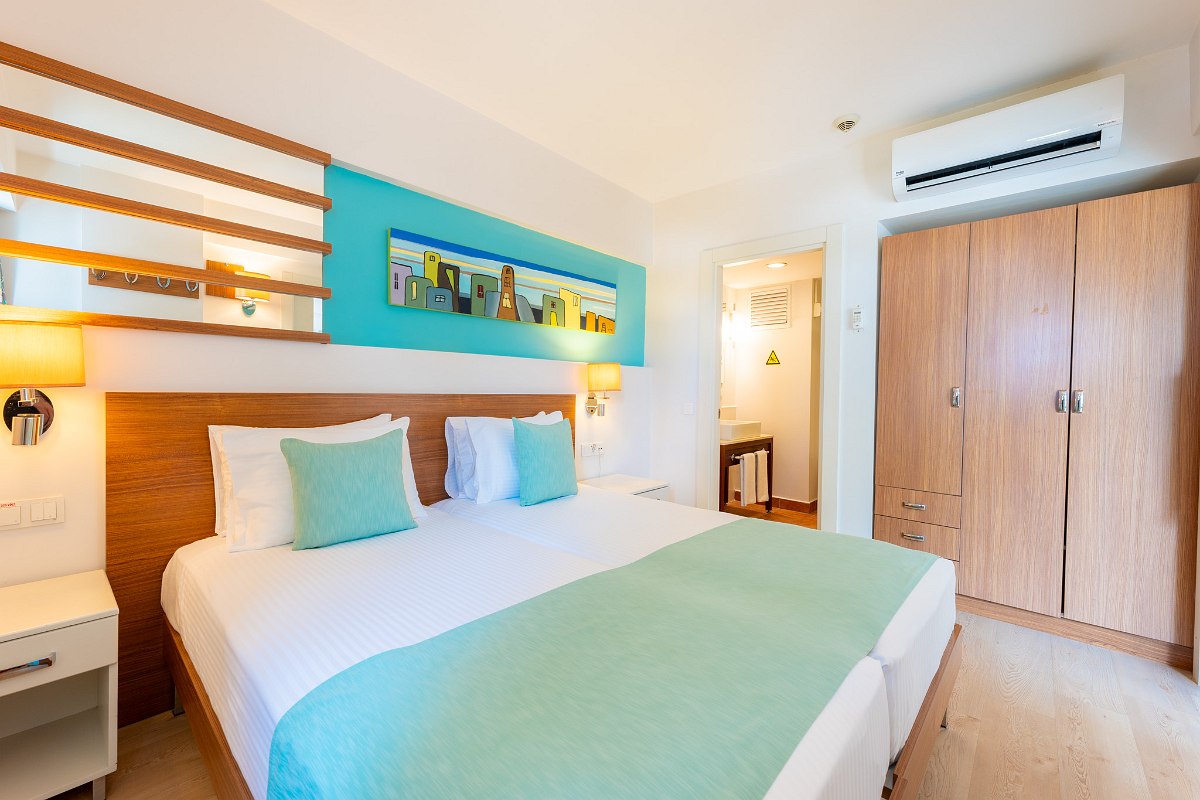 Cozy hotel room with twin beds, green bedding, a wooden headboard, and wall-mounted lights. The room opens to a living area with a sofa and kitchen. There's an air conditioner above the wardrobe and a bathroom visible through an open door.