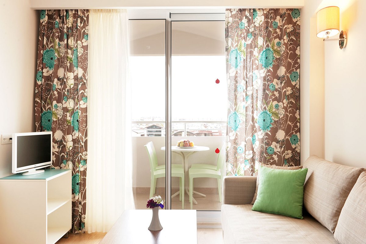 Cozy living room with a beige sofa and green pillow, a white TV stand with a small TV, floral curtains, and a balcony with a table and chairs outside.