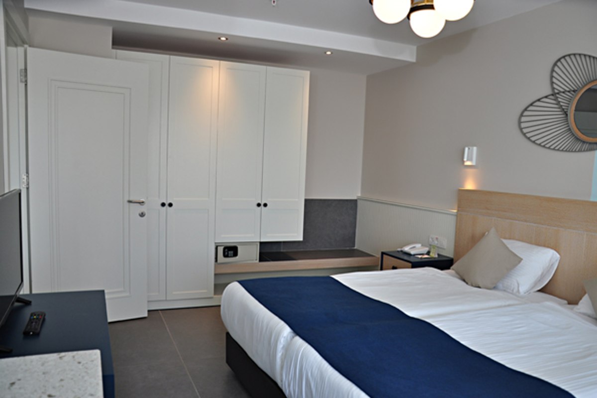 Simple, clean hotel room with twin beds pushed together, covered in white bedding with dark blue runners. The room features a large white wardrobe, a black TV stand with a TV, a nightstand, and a gray cushioned bench near the wardrobe. The color scheme is neutral with light walls and dark floors.