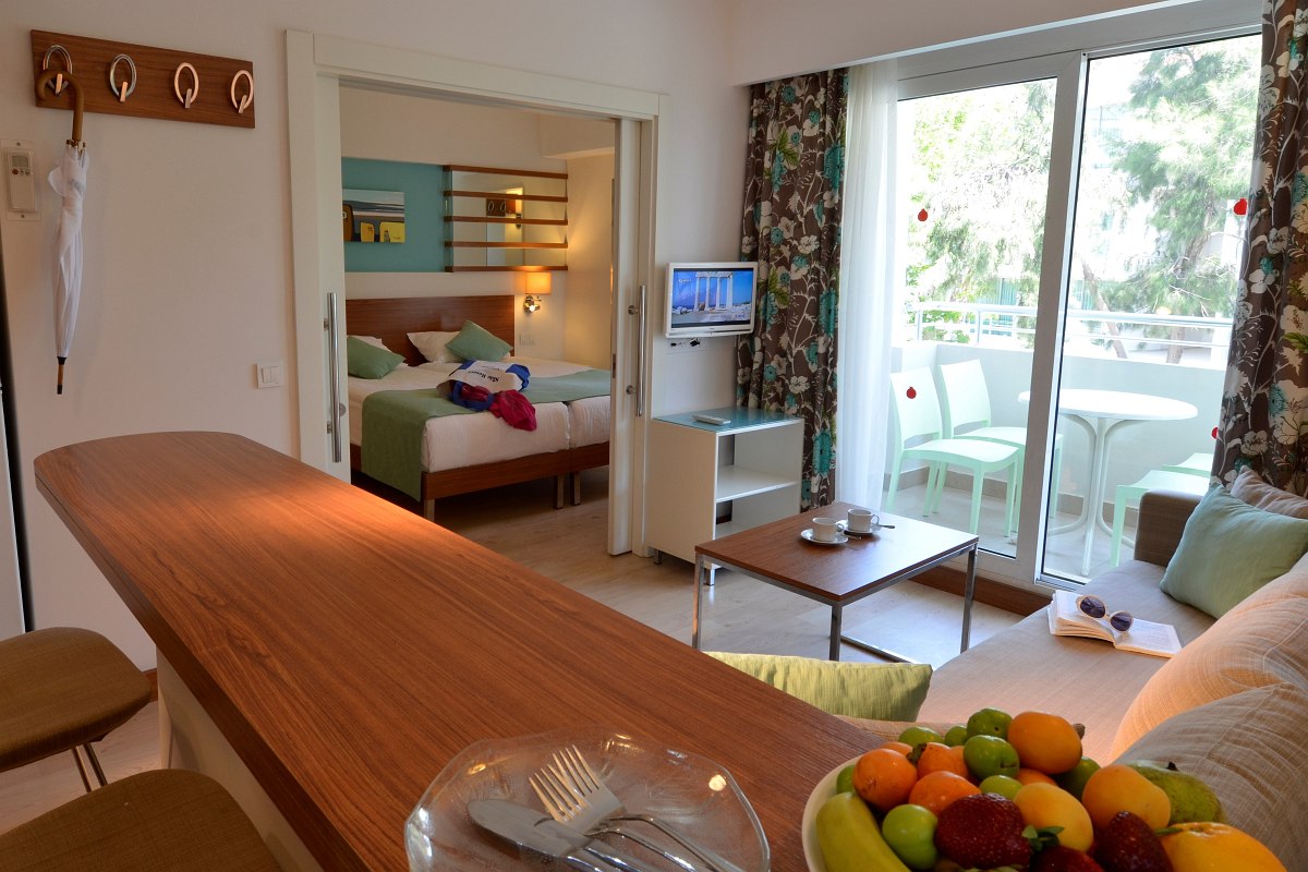 Cozy, modern suite with a small kitchen counter and bar stools. The living area has a light sofa with colorful cushions, a coffee table, and a flat-screen TV. Through the sliding glass door, there's a balcony with a small table and two chairs overlooking green trees. The bedroom, visible through a sliding door, has a bed with green and white bedding and bedside lamps. Bright and inviting with a fresh atmosphere.
