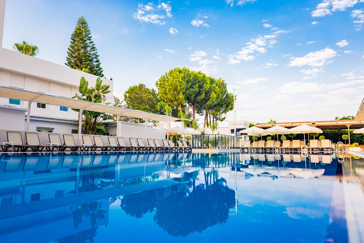 Outdoor pool with blue sun loungers and umbrellas, next to a basketball court. Sunny atmosphere.