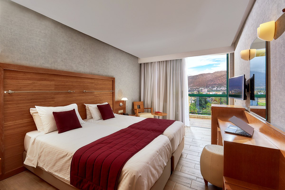 A modern hotel room with a large bed dressed in white linens and red accents, wooden furniture, a wall-mounted TV, and a balcony offering a scenic mountain view.