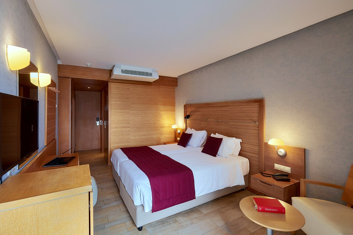 A cozy hotel room with a large bed, red accents, wooden furniture, a wall-mounted TV, and soft lighting creating a warm atmosphere.
