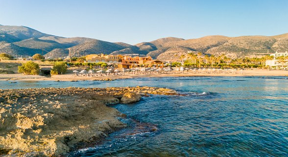 Coastal landscape with sandy beach, sun loungers, hotel complex, palm trees and mountains in the background at sunset.