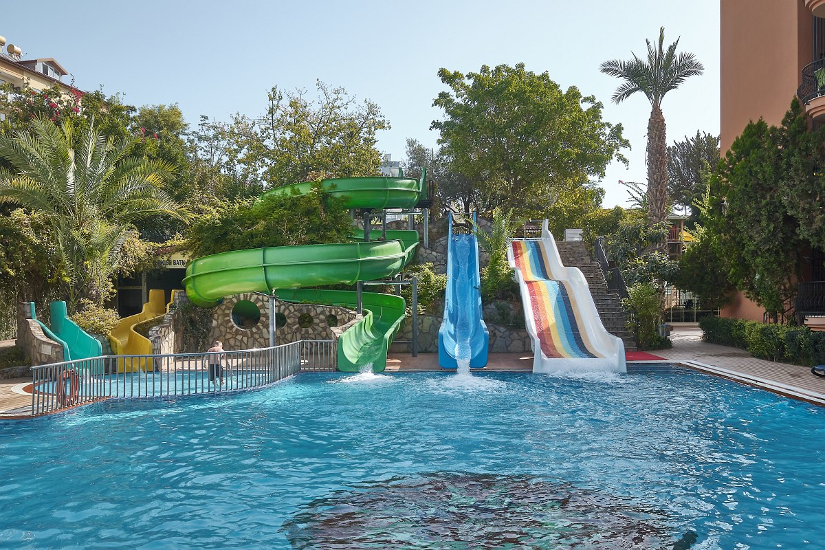 Pool with colourful water slides and blue deckchairs at the edge of the pool, next to a modern, bright hotel building.