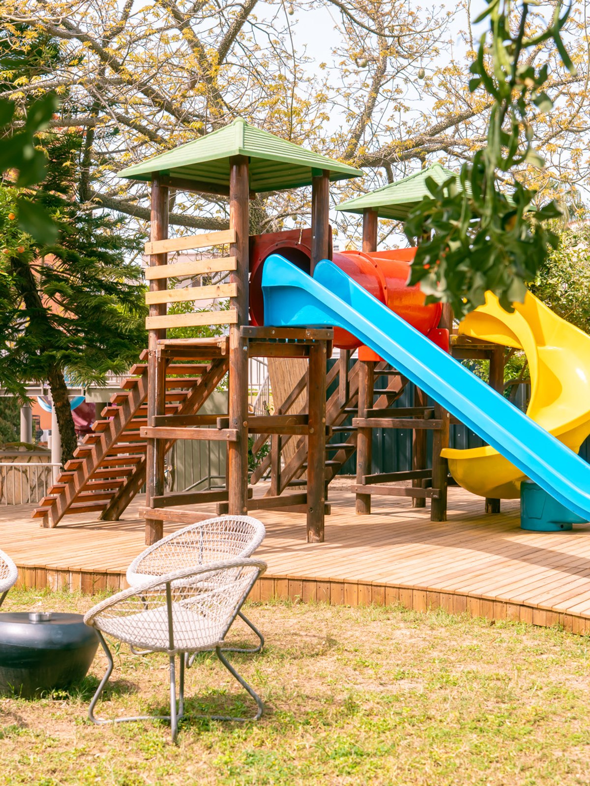 A white building with large windows that houses the ‘CALIMIGO KIDS CLUB’. A tree wrapped in a colourful crochet pattern stands next to it, surrounded by green plants.