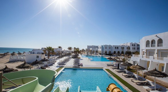 Large pool with loungers, palm trees, and a view of the bright main hotel building in sunshine