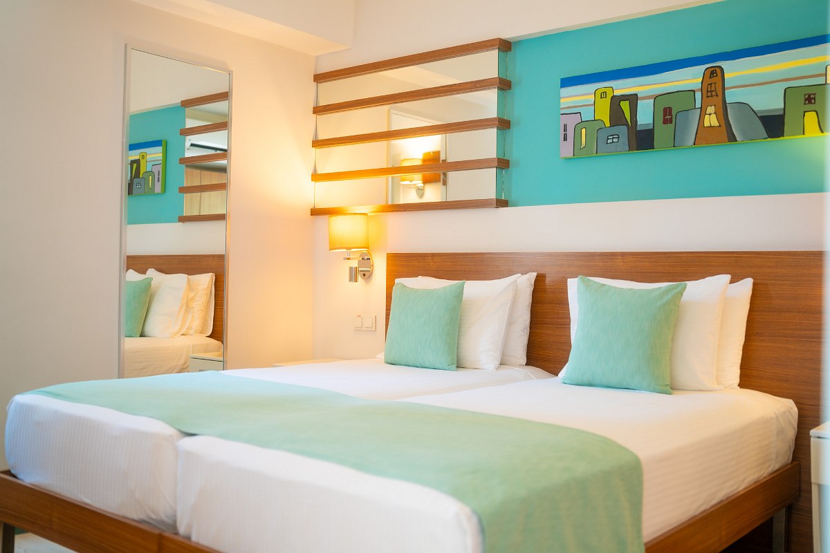 Chambre lumineuse avec deux lits simples, coussins verts, table de chevet, penderie ouverte et décoration murale colorée.
