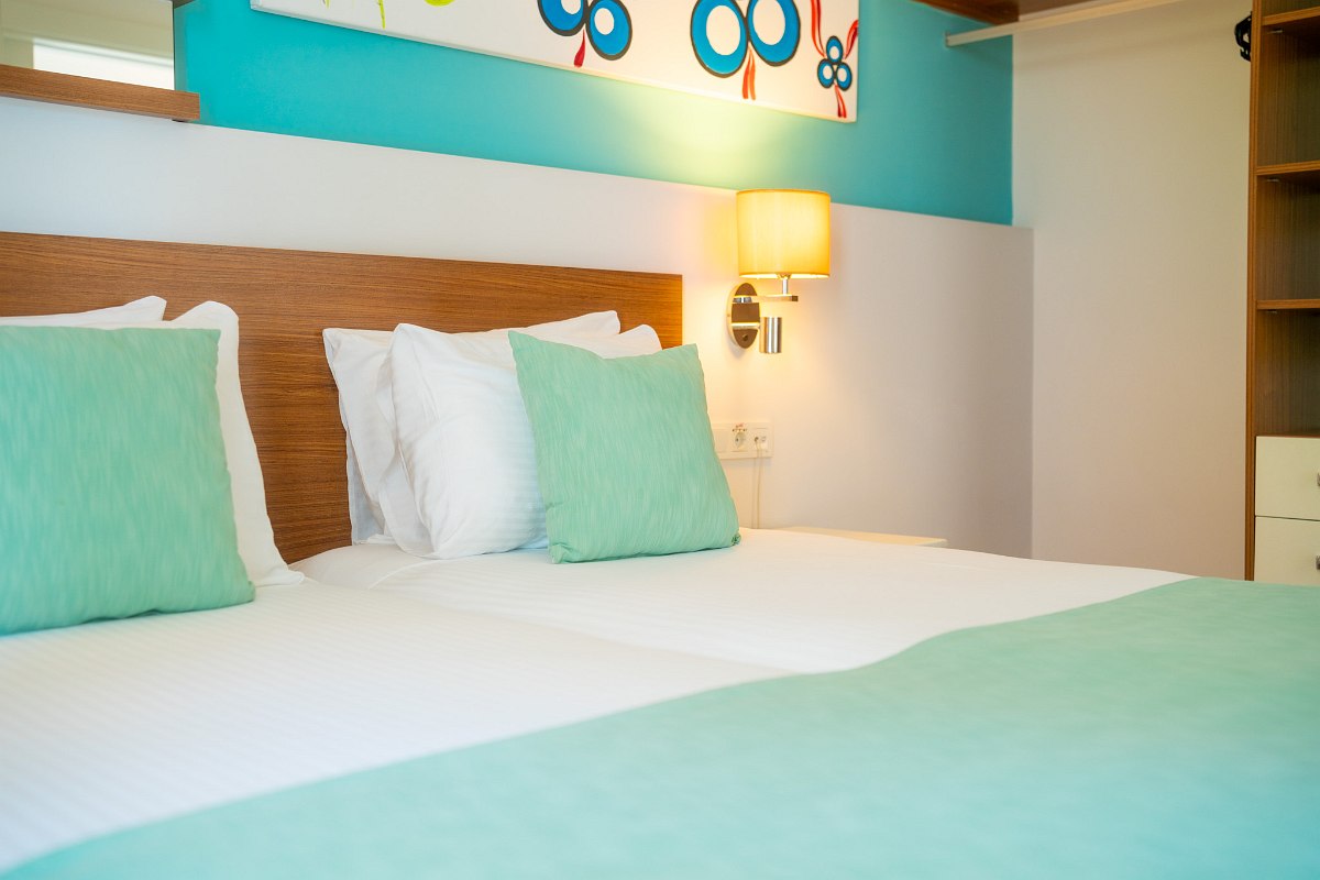 Chambre moderne avec de grandes armoires blanches, un lit double avec une couette bleu foncé et une télévision posée sur une table noire. Éclairage discret et miroir au mur.