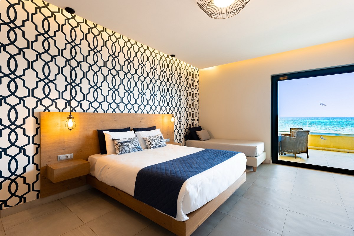 Chambre d'hôtel moderne avec lit double, canapé, bureau, vue sur la mer et balcon avec siège.
