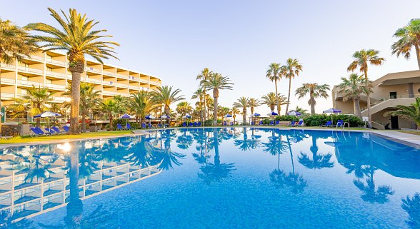 Grande piscine d'hôtel avec palmiers et chaises longues, en arrière-plan, bâtiment d'hôtel à plusieurs étages par temps clair.